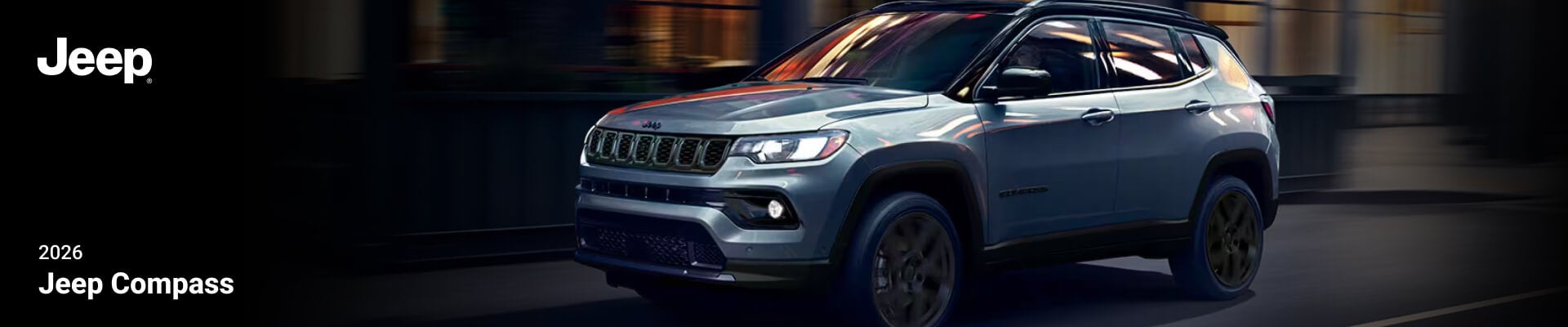 A silver 2026 Jeep Compass driving down a city street at night.
