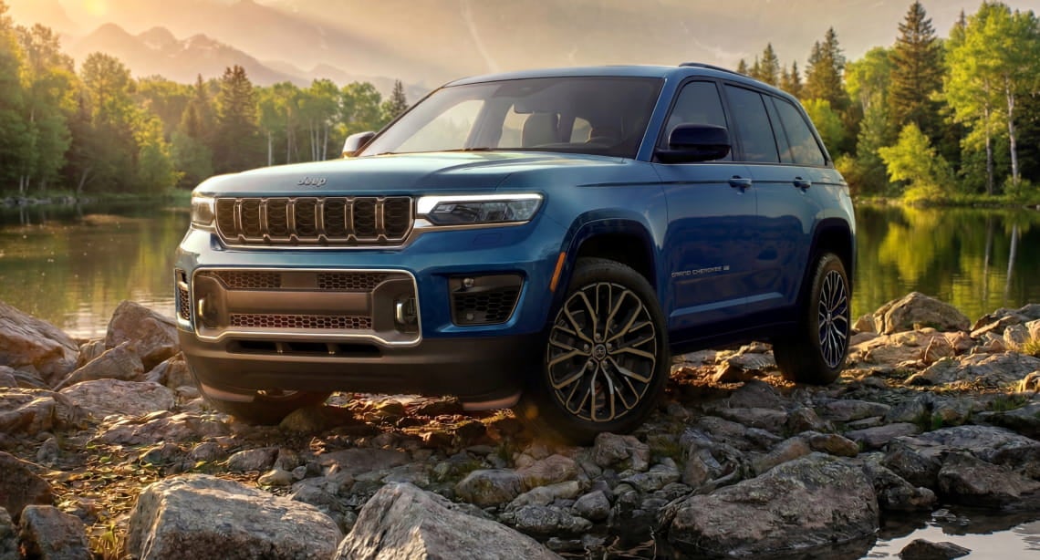 Blue Jeep Grand Cherokee shown from a front three-quarter angle on rocky terrain beside a lake.