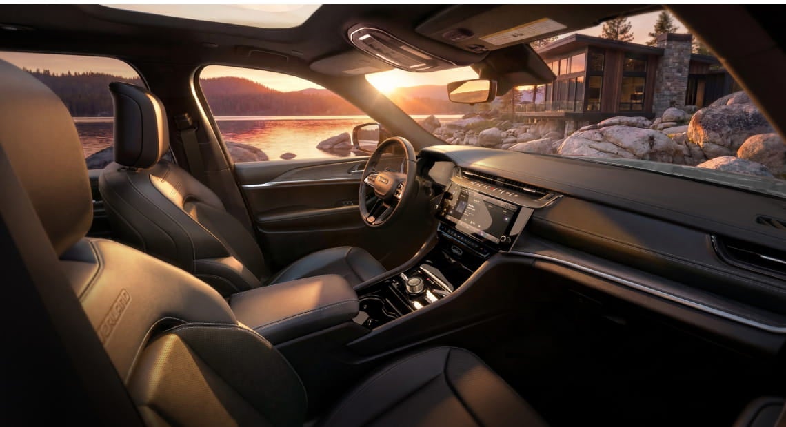 Jeep Grand Cherokee interior with black seating, a center touchscreen, panoramic roof, and a view of a lakeside setting at sunset.